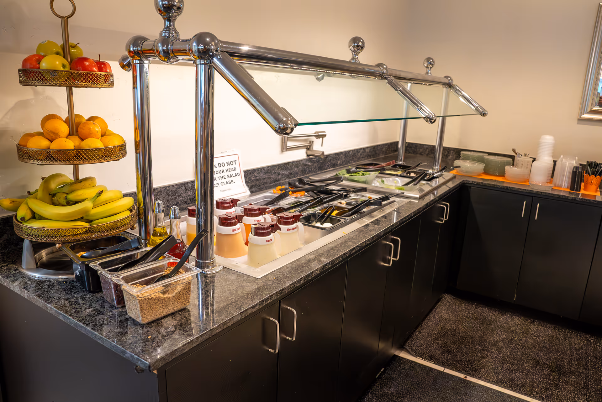 A salad bar station with various fresh fruits including apples, oranges, and bananas on a tiered stand, alongside containers of salad toppings and dressings under a glass sneeze guard in a senior living facility.