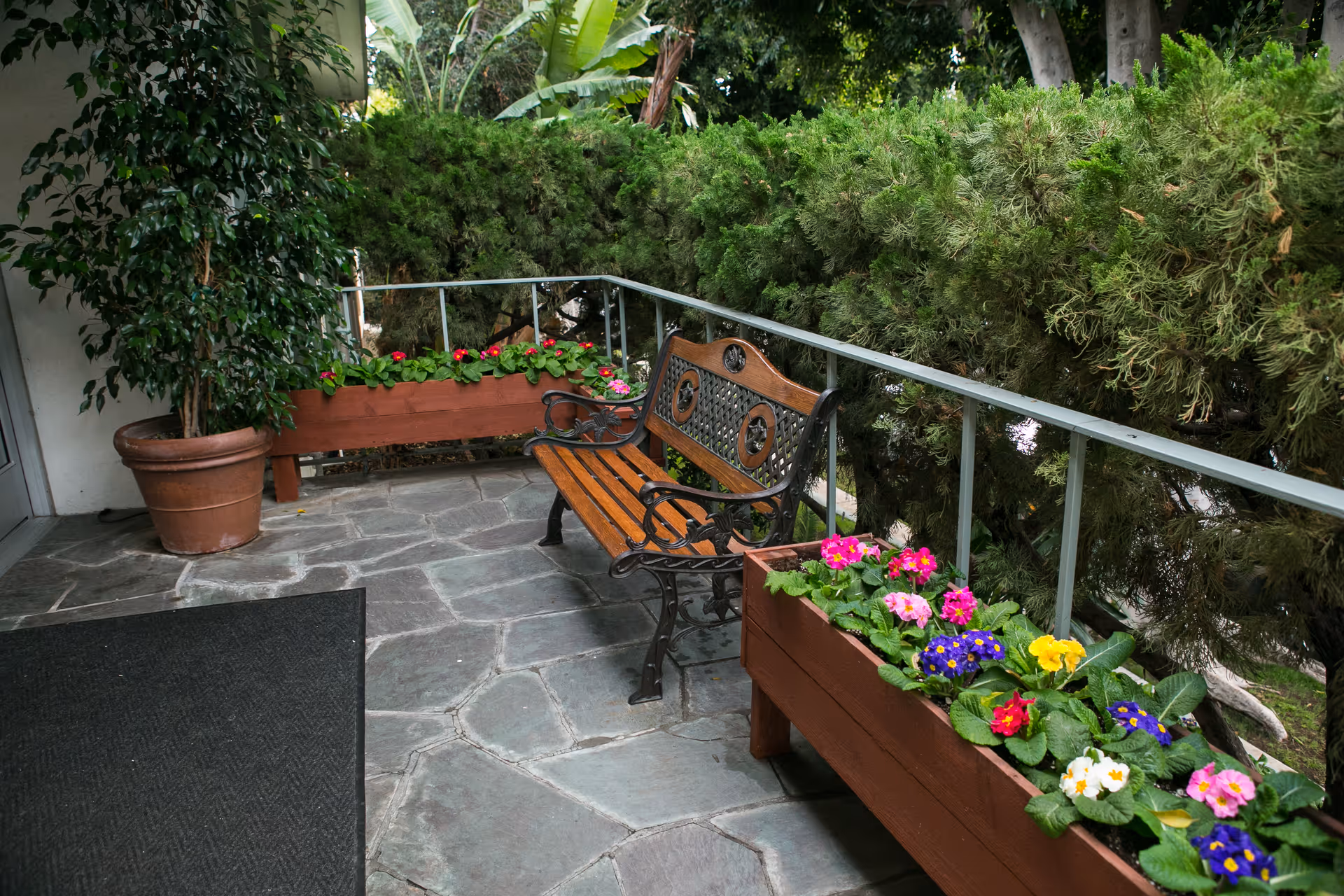 Outdoor patio area with a wooden bench featuring decorative metal armrests and backrest, surrounded by green shrubs and colorful flowers planted in wooden planters. A large potted plant is placed near the entrance door on a stone-tiled floor.