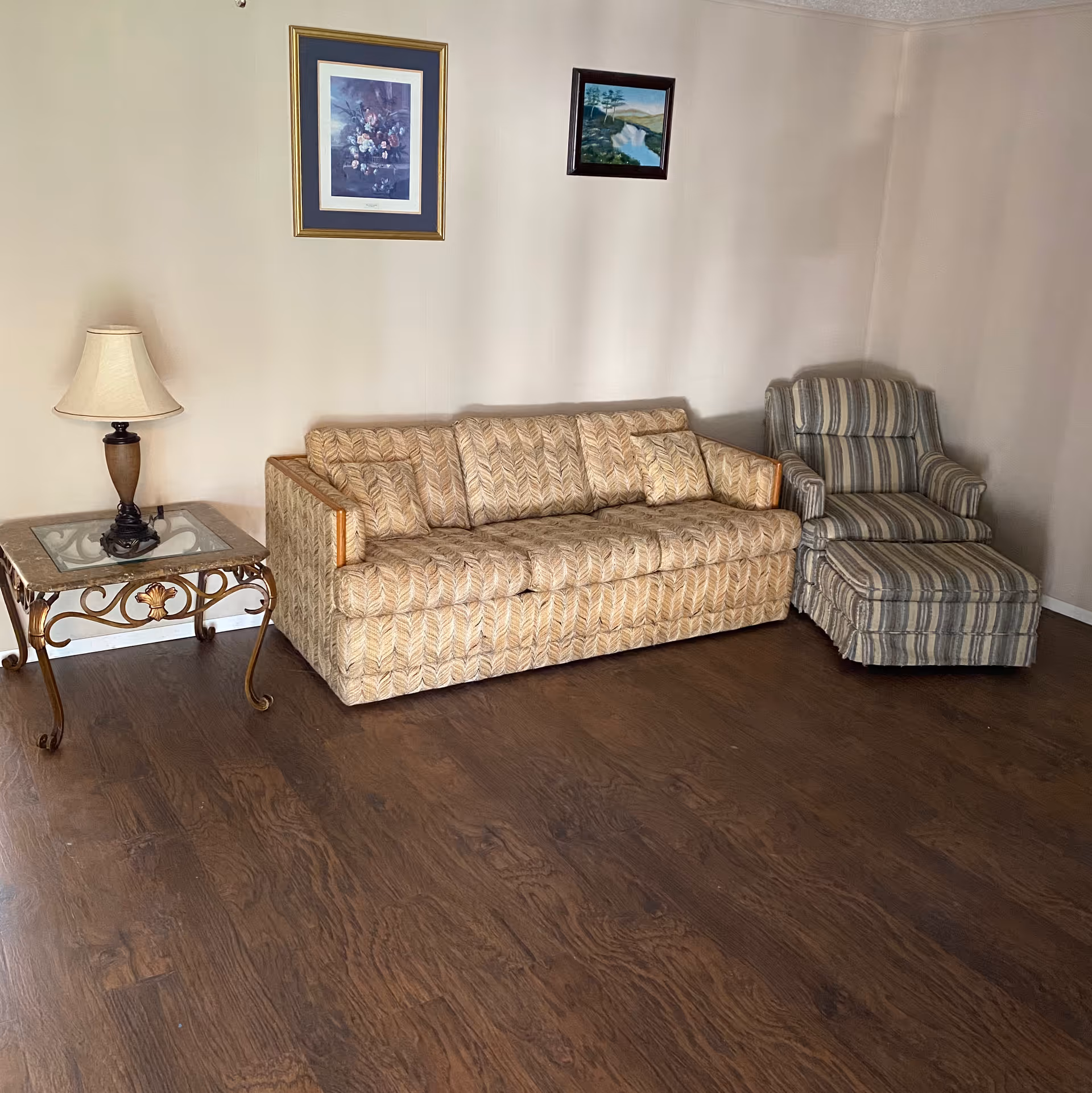 A living room area with a patterned beige sofa, a striped armchair with a matching ottoman, a glass-top side table with ornate metal legs, and a table lamp. Two framed paintings hang on the beige wall above the sofa.