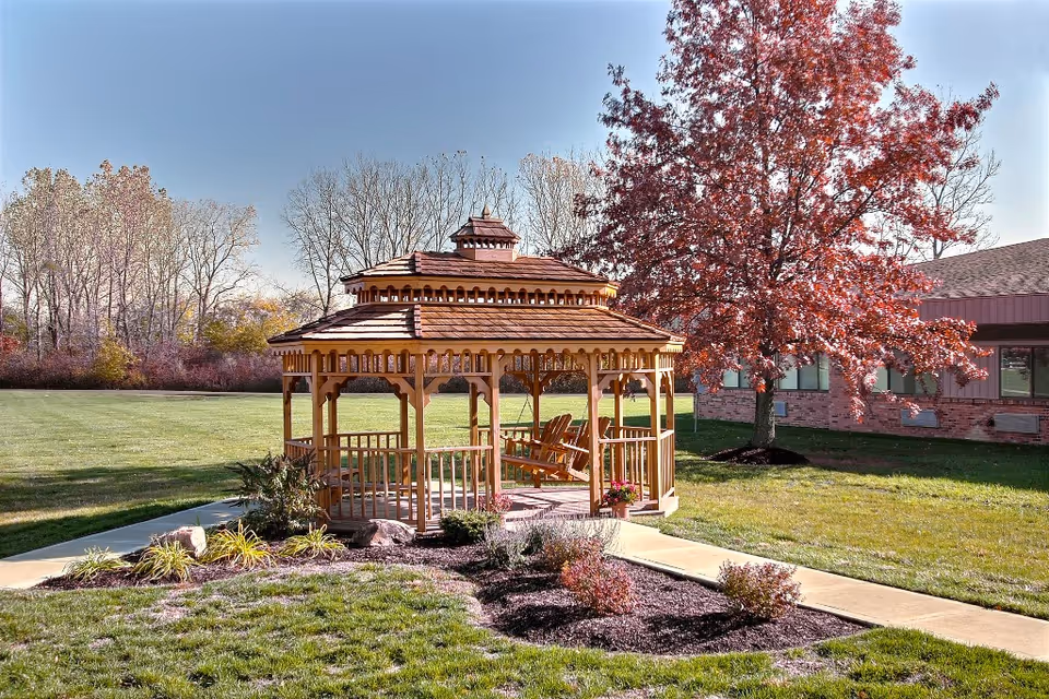 A wooden gazebo on a manicured lawn with chairs, a red-leaved tree, and a brick building in the background.