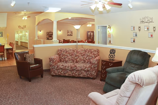 A cozy living room area with a floral patterned sofa, a dark green recliner, a brown leather chair, and a light pink recliner. There is a wooden side table with an owl decoration, a floor lamp, and framed pictures on the wall. The room has beige walls, carpeted floor, and ceiling fans with lights. In the background, there is a dining area with tables and chairs.