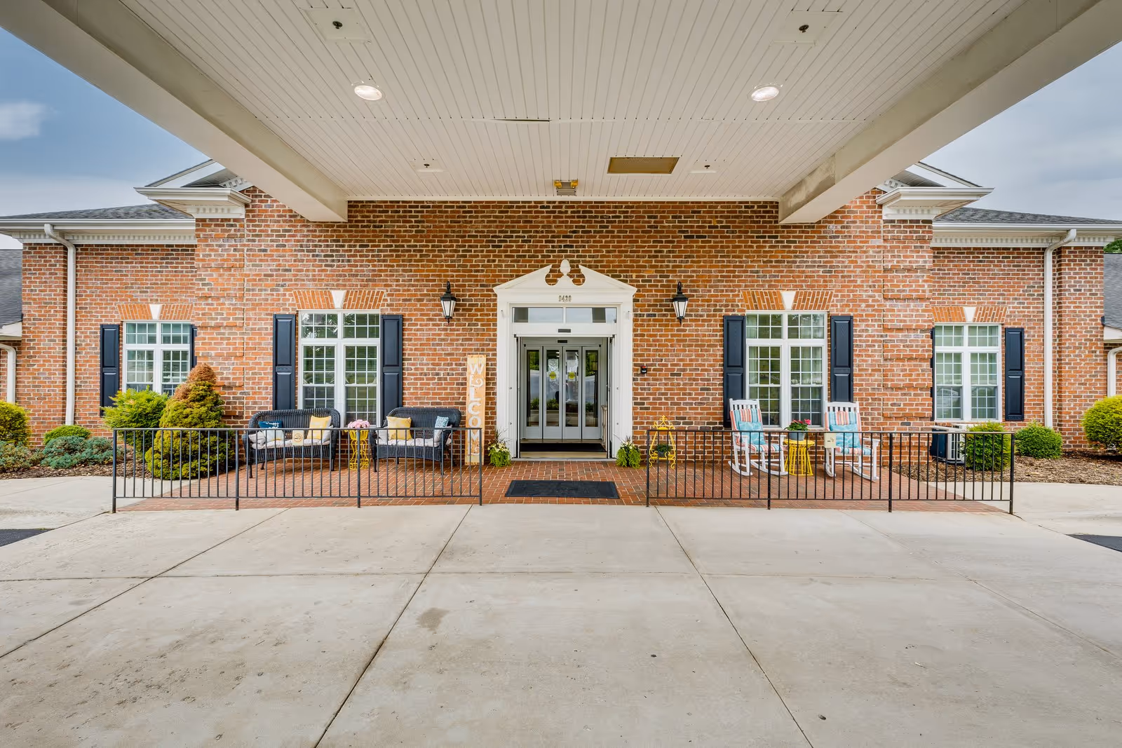 Front entrance of Mallard Ridge Assisted Living facility featuring a brick exterior with white trim, two black lantern-style wall lights, and double glass doors. There are two seating areas on either side of the entrance with black and white chairs, small yellow tables, and decorative pillows. A vertical wooden welcome sign is placed near the left seating area. The entrance is covered by a white ceiling with recessed lighting.