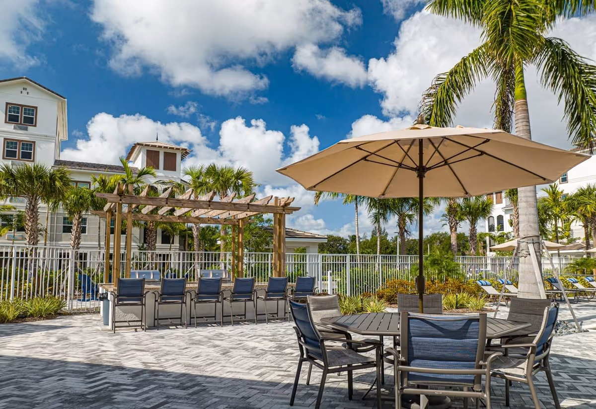 Outdoor patio area at Arcadia Gardens with a round table and chairs under a large beige umbrella, a pergola with bar seating and grills, palm trees, and a multi-story building in the background under a partly cloudy blue sky.