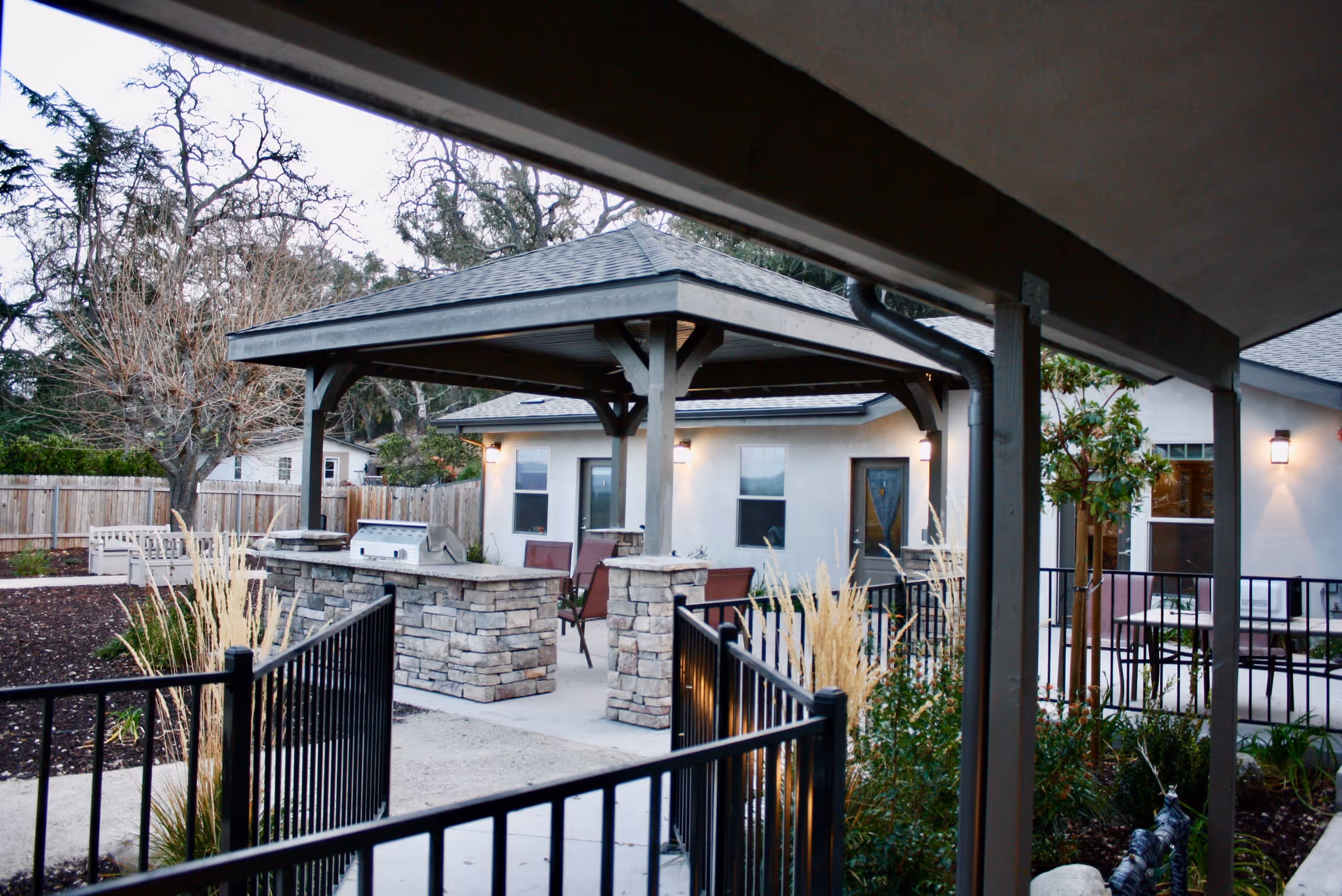 Covered pavilion with a stone grill island and outdoor seating on a courtyard patio next to the facility building.