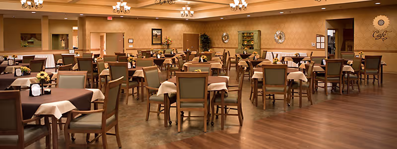 A spacious dining room with multiple wooden tables and chairs arranged neatly. Each table is covered with a brown tablecloth and a white napkin, and decorated with small flower arrangements. The room has warm lighting with chandeliers on the ceiling and a wooden floor. On the far wall, there is a sign that reads 'Cafe' with decorative mirrors and shelves.