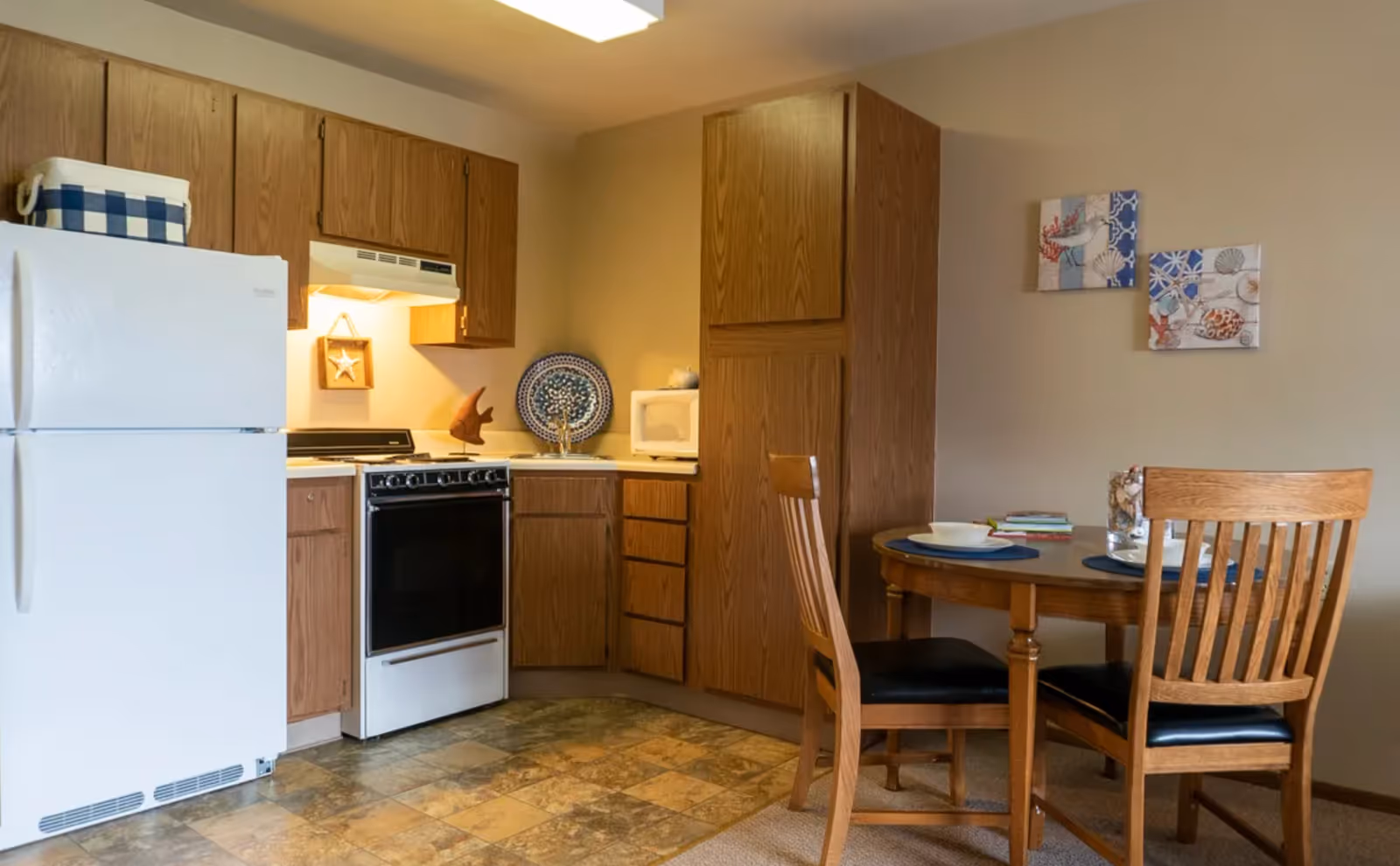 A small kitchen area with wooden cabinets, a white refrigerator, a white stove with an overhead vent, a microwave on the countertop, and a round wooden dining table with two chairs. The wall has two decorative paintings with a coastal theme.