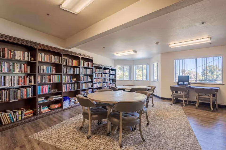 A bright room with large windows featuring a library with multiple bookshelves filled with books along one wall. In the center, there are round tables surrounded by cushioned chairs on wheels. Against the windows, there are two desks with chairs and a computer on one desk. The floor is a combination of wood and a large area rug.
