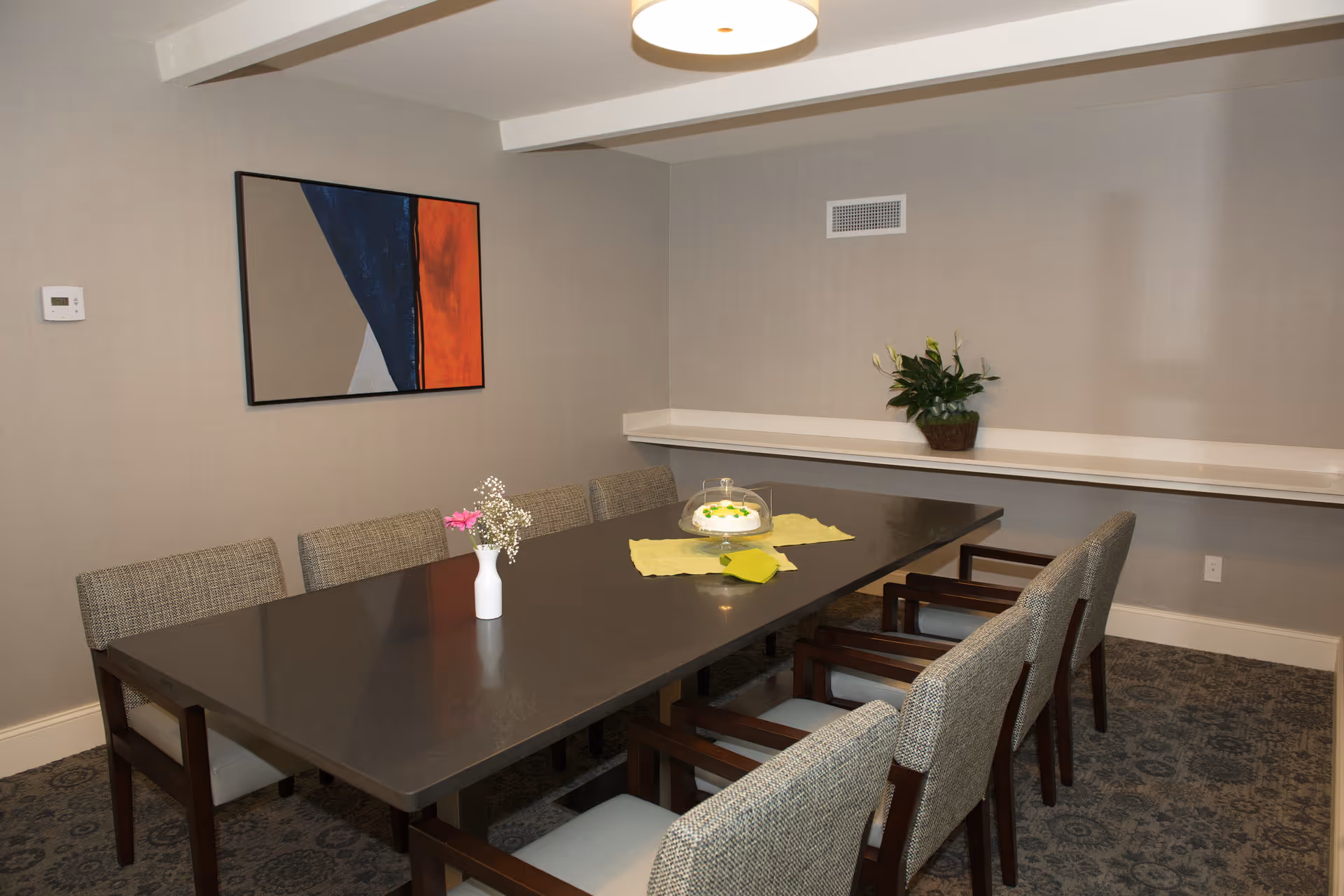 A dining room with a long rectangular table surrounded by eight upholstered chairs. On the table, there is a small white vase with pink and white flowers, a cake under a glass cover, and a yellow cloth. The walls are light gray with a modern abstract painting hanging on one side and a potted plant on a narrow shelf along another wall. The room has a patterned carpet and a ceiling light fixture.