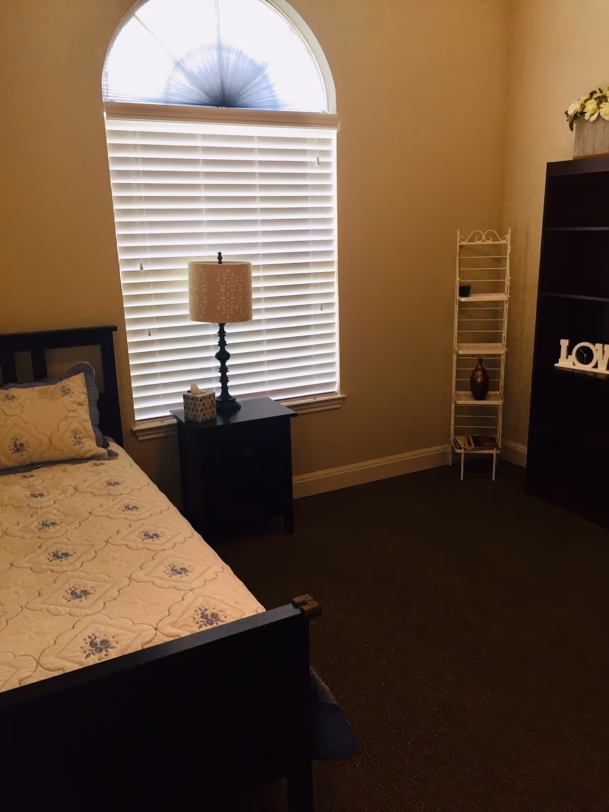 A bedroom with a single bed featuring a floral quilt and pillow, a black nightstand with a decorative lamp and tissue box, a window with white blinds and an arched top, a white metal shelf with decorative items, and a dark wooden bookshelf with a 'LOVE' sign and a flower arrangement on top.