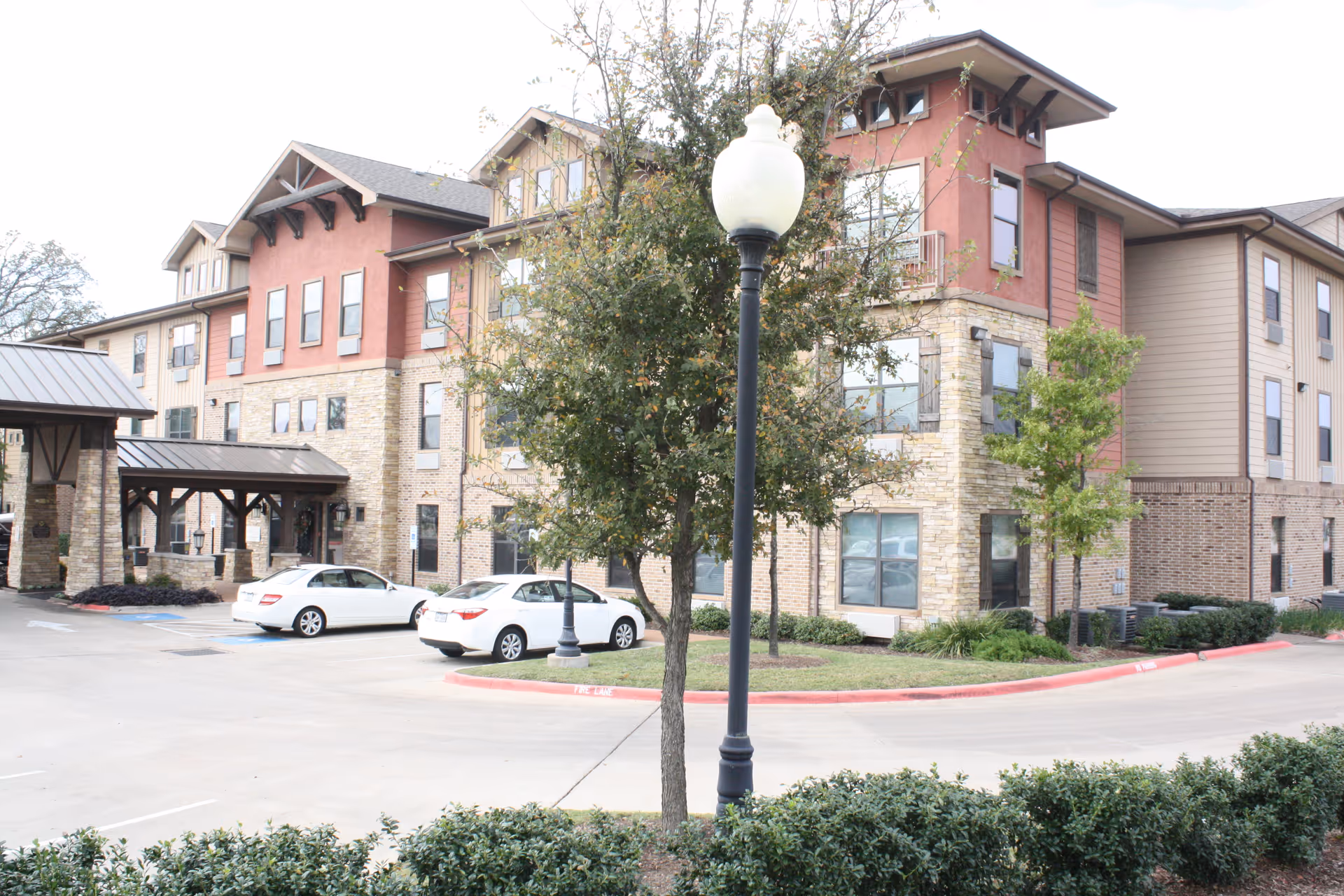 Exterior view of a multi-story senior living facility building with a mix of stone and siding facade. There are several windows, a covered entrance, a parking lot with two white cars, a street lamp, and some trees and shrubs in the landscaped area.
