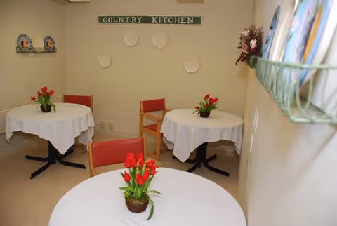 Small dining area with three round tables covered with white tablecloths, each table decorated with a small pot of red tulips. The walls are beige, with decorative plates mounted on one wall and a sign that reads 'COUNTRY KITCHEN'. There are red and wooden chairs around the tables.