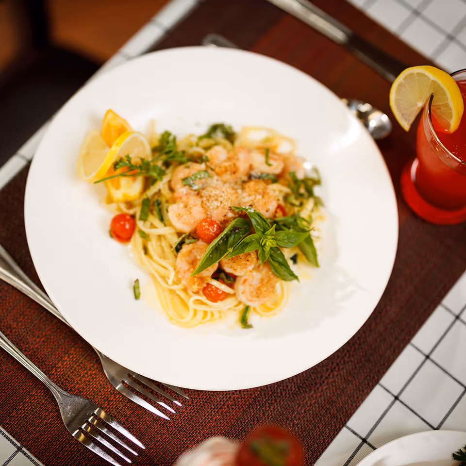 A white plate with a serving of shrimp pasta garnished with fresh basil, cherry tomatoes, and lemon wedges. The plate is set on a brown placemat with a fork and knife beside it. A glass of red beverage with a lemon slice on the rim is also visible.