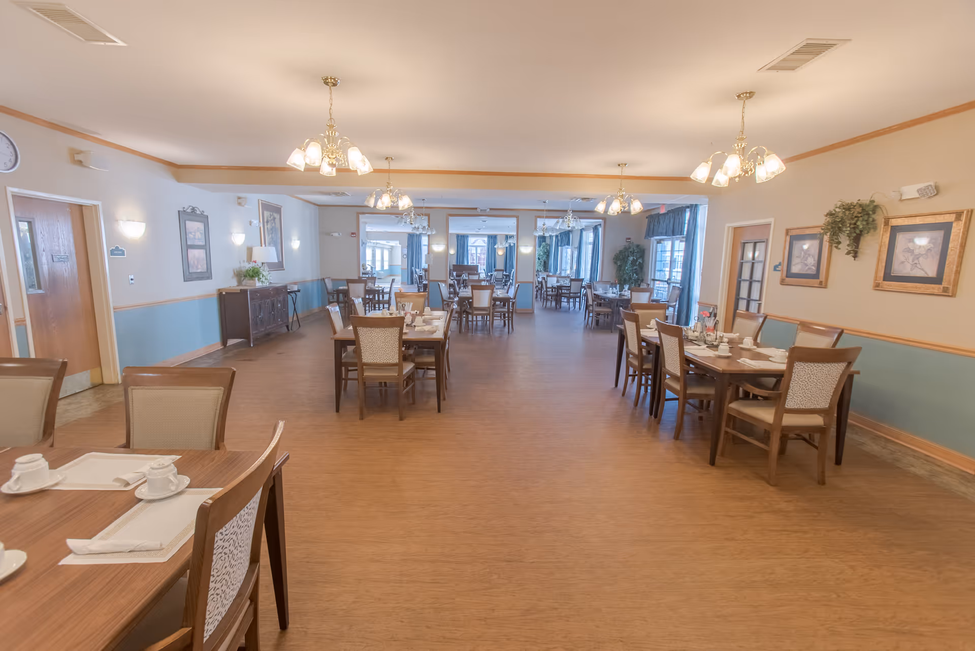 A spacious dining room in a senior living facility with multiple wooden tables and chairs arranged neatly. Each table is set with white cups, saucers, and napkins. The room has warm lighting from chandeliers hanging from the ceiling, light blue and beige walls with framed artwork, and large windows with curtains allowing natural light to enter.