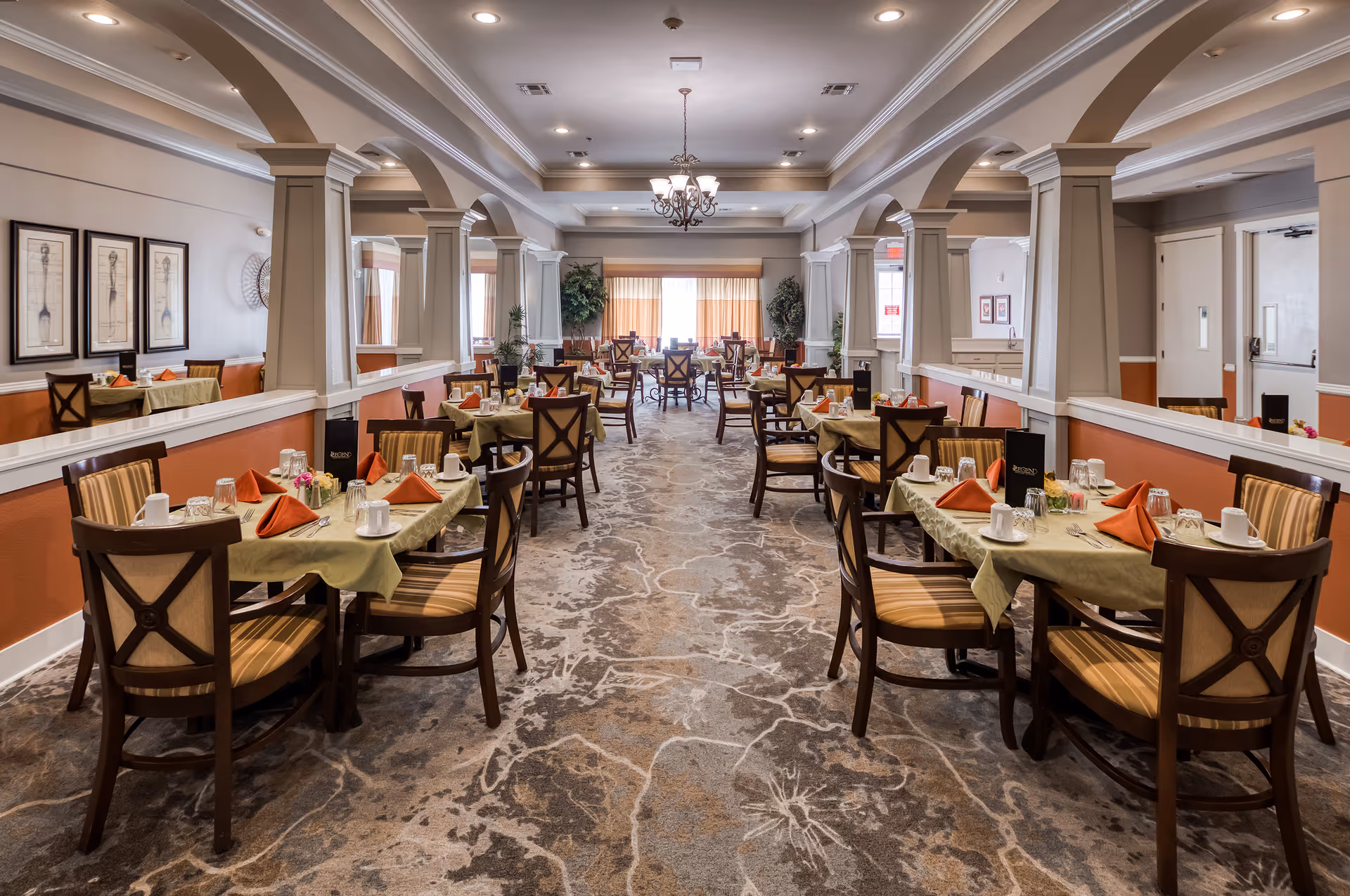 A spacious dining room with multiple tables set for meals, featuring green tablecloths, orange folded napkins, cups, glasses, and silverware. The room has a patterned carpet, arched columns, framed artwork on the walls, and a chandelier hanging from the ceiling. Large windows with curtains allow natural light to fill the space.