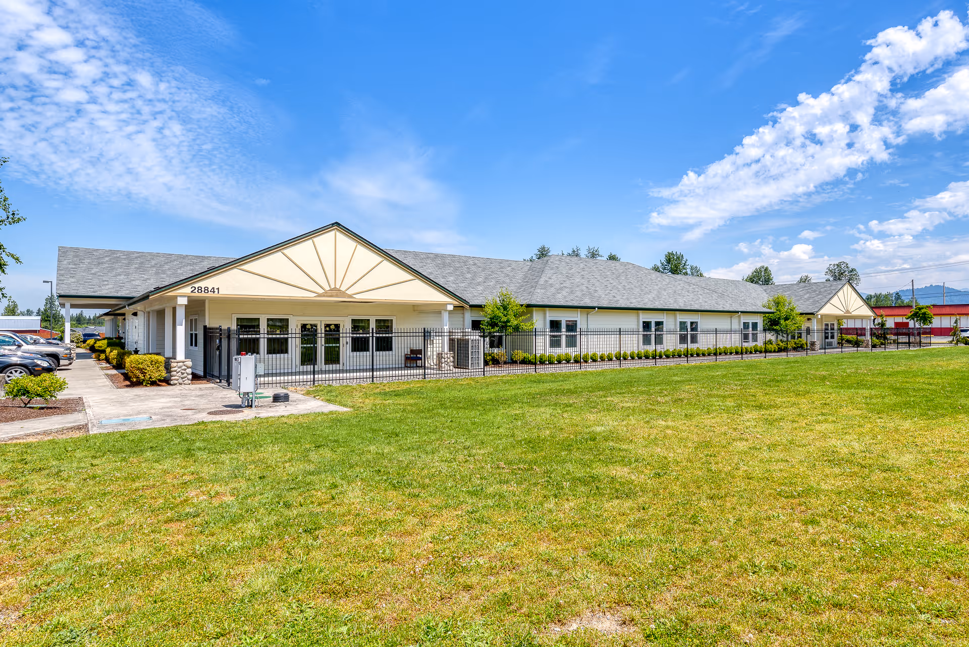 Exterior view of a single-story assisted living and memory care facility building with a gray roof, cream-colored walls, and a fenced patio area. The building is surrounded by a well-maintained lawn and landscaping under a partly cloudy blue sky.