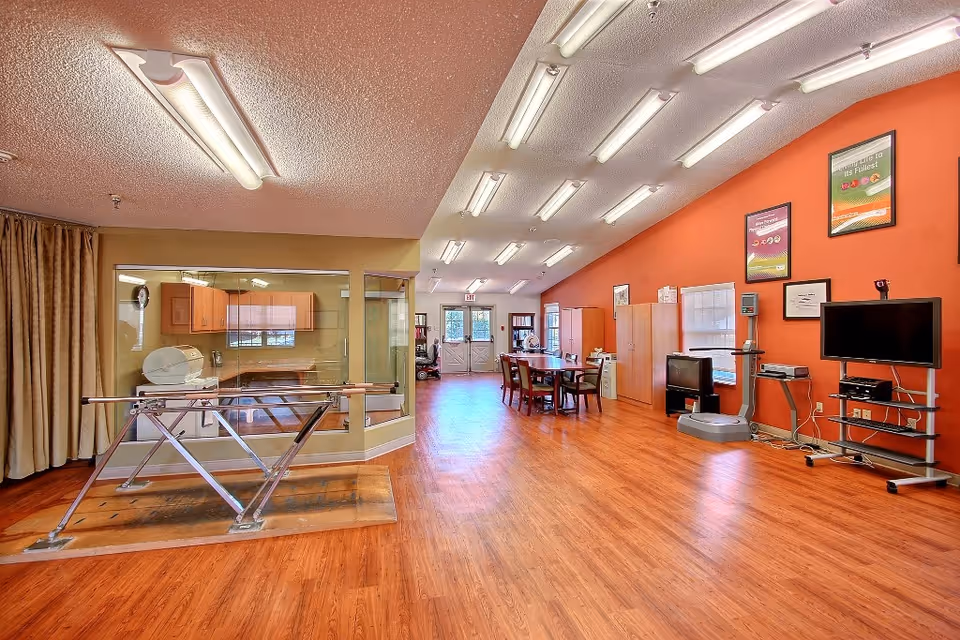 A spacious room with wooden flooring and bright lighting, featuring a parallel bar setup for physical therapy on the left, a table with chairs in the center, and various exercise and entertainment equipment along the right wall. The walls are painted in warm tones with framed posters, and there are windows and a door at the far end.