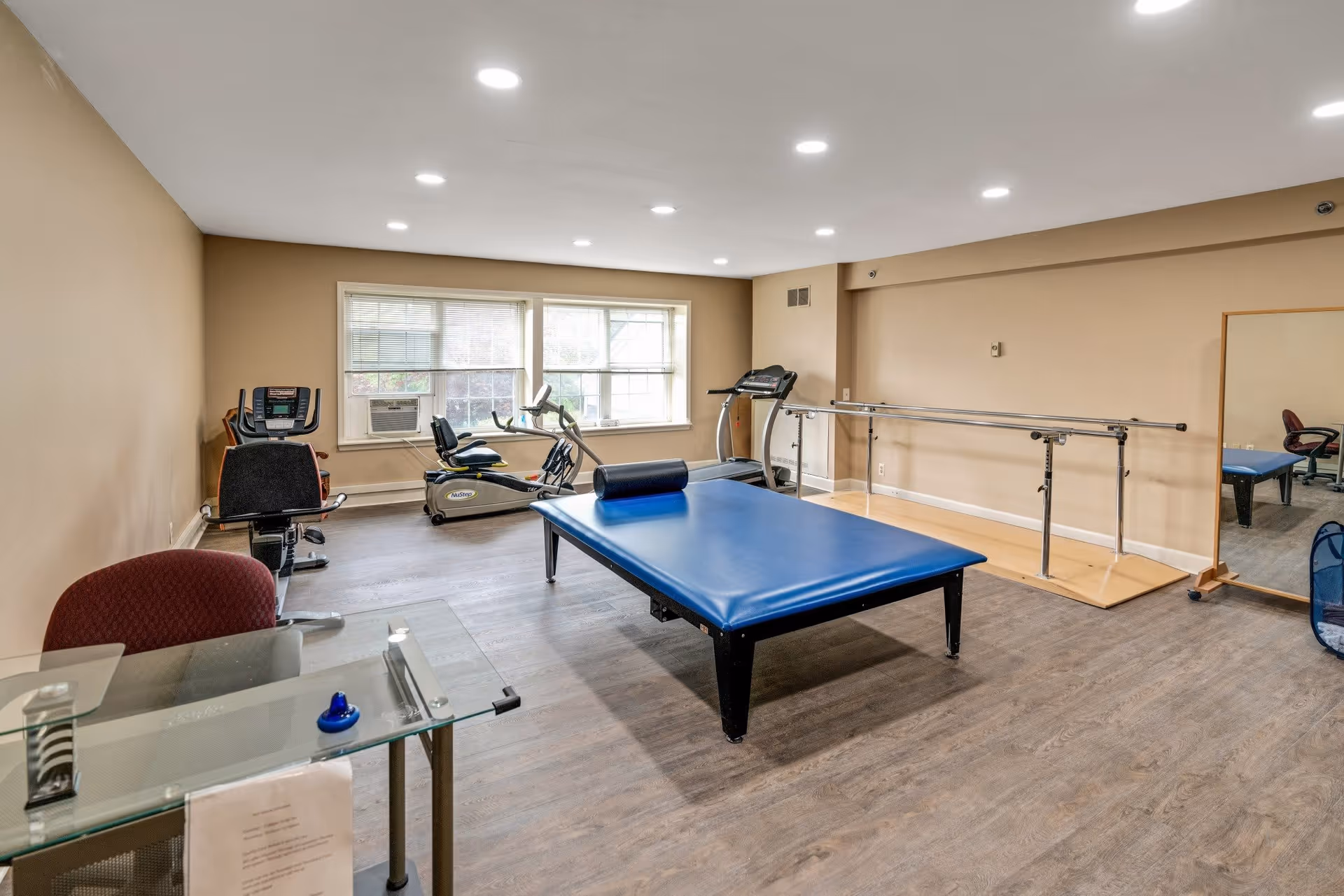 A rehabilitation or physical therapy room with exercise equipment including a recumbent bike, treadmill, and a large blue padded therapy table. There is a ballet barre along one wall and a large mirror next to it. The room has beige walls, wood flooring, and a window letting in natural light. A glass desk with a red chair is visible in the foreground.