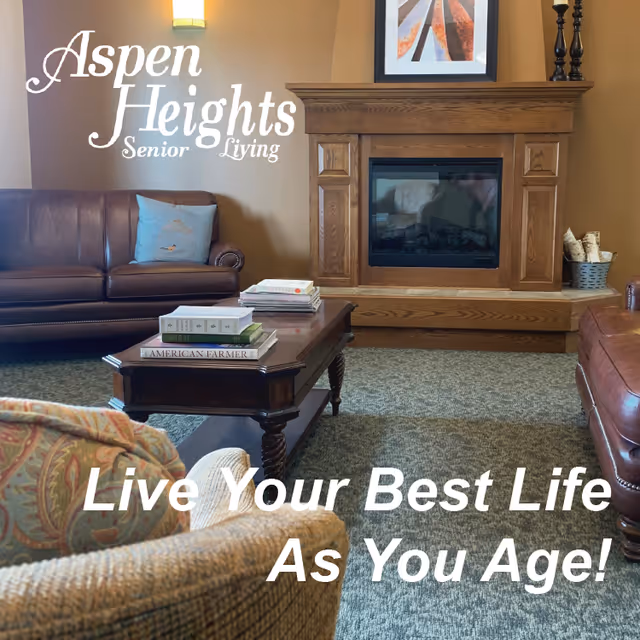 A cozy senior living room with brown leather sofas, a wooden coffee table stacked with books, a fireplace with a wooden mantel, and a framed picture above it. The room has warm lighting and a carpeted floor.