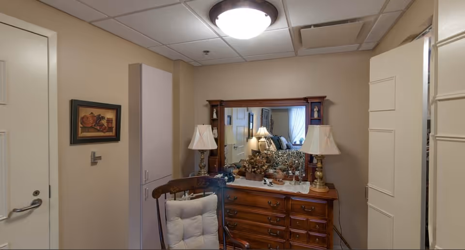 Interior view of a room featuring a wooden dresser with a large mirror, two table lamps, decorative items, and a cushioned wooden chair. The walls are beige, and there is a framed picture on the left wall. A door is partially open on the right side.