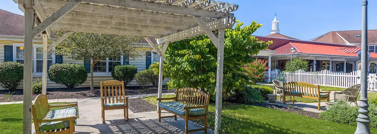Outdoor seating area at Milford Place featuring wooden benches and chairs with cushions under a white pergola, surrounded by green grass, bushes, and trees with a building and white picket fence in the background.