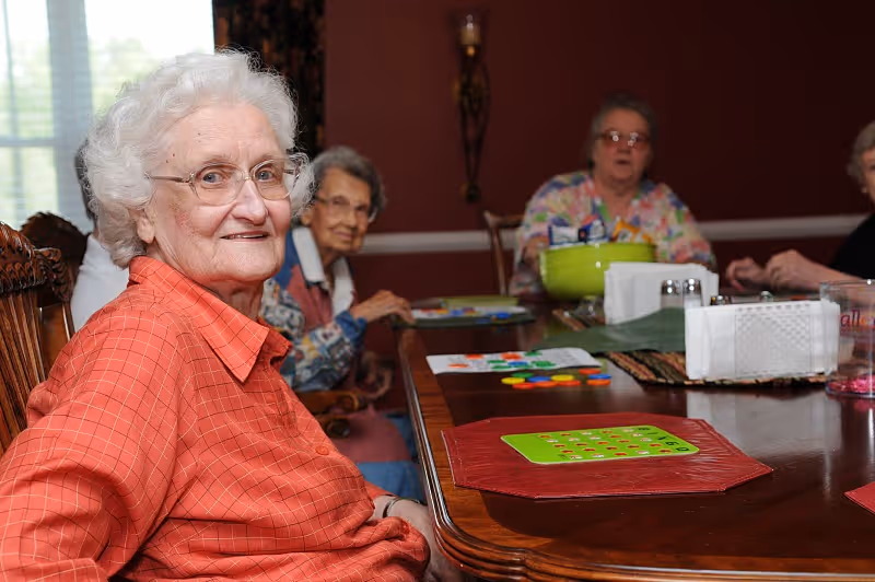 A group of elderly women sitting around a wooden table in a room with dark red walls, engaging in a board game or activity. One woman in an orange shirt is smiling at the camera, while others are focused on the game pieces on the table.