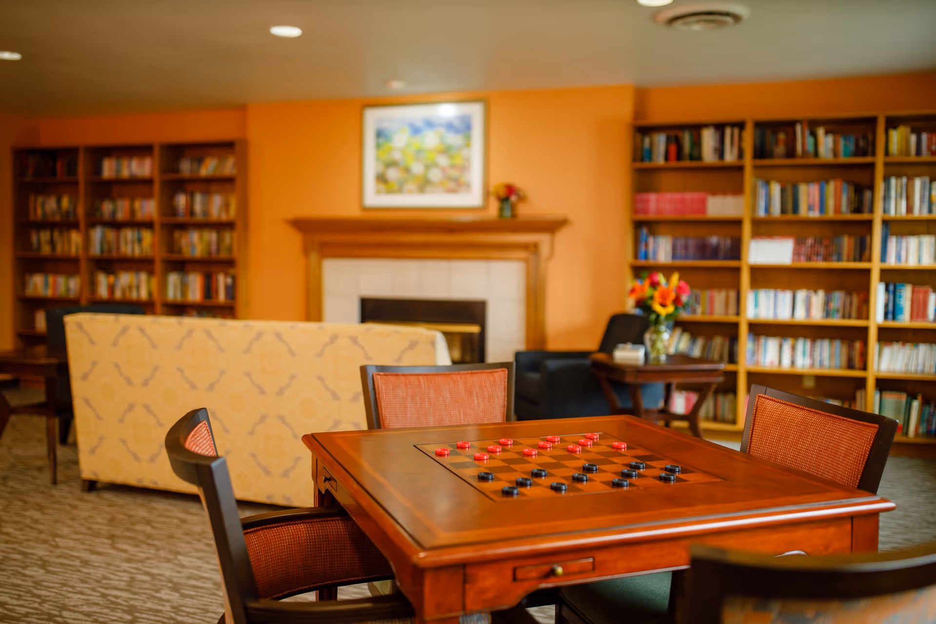 Communal living room with a wooden game table set for checkers, upholstered chairs and sofas, bookshelves and a fireplace.