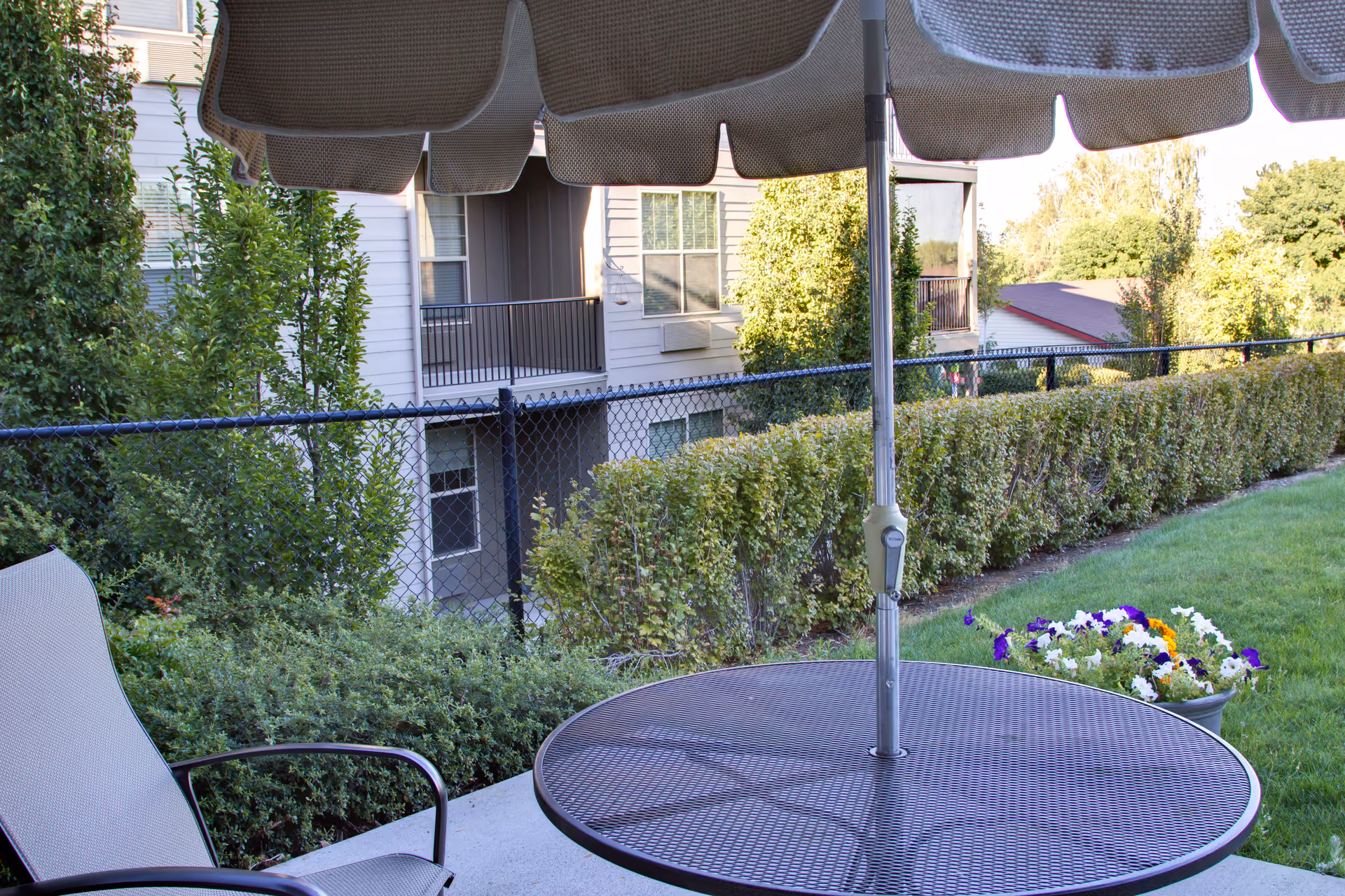 Outdoor patio area with a round metal table and umbrella, a chair, green bushes, a flower pot with colorful flowers, and a view of a building and trees in the background.