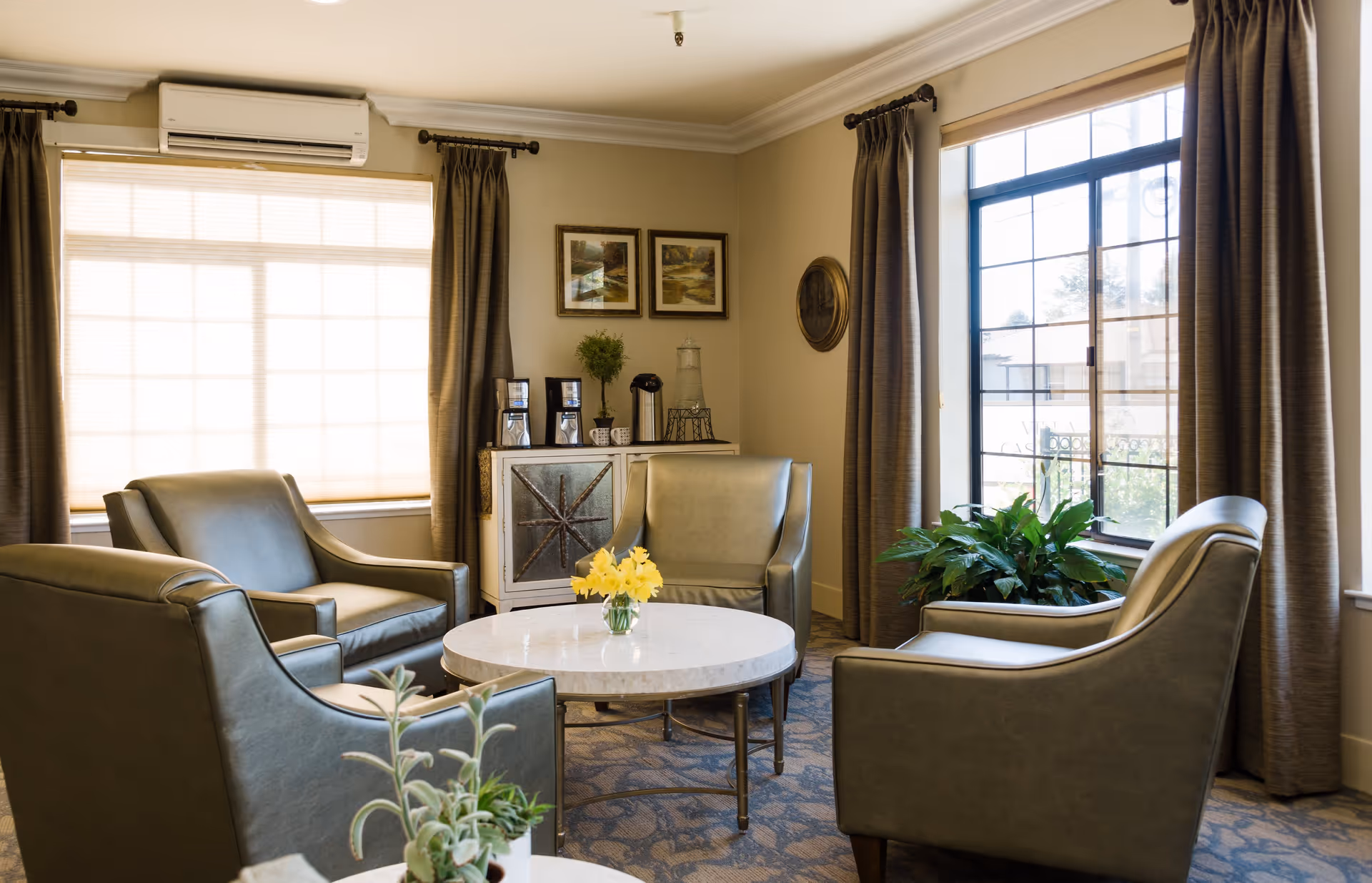 A cozy sitting area in an assisted living facility with four green armchairs arranged around a round white marble table with a small vase of yellow flowers. The room has large windows with brown curtains, a cabinet with coffee makers and cups, framed artwork on the walls, and a potted plant near the window.