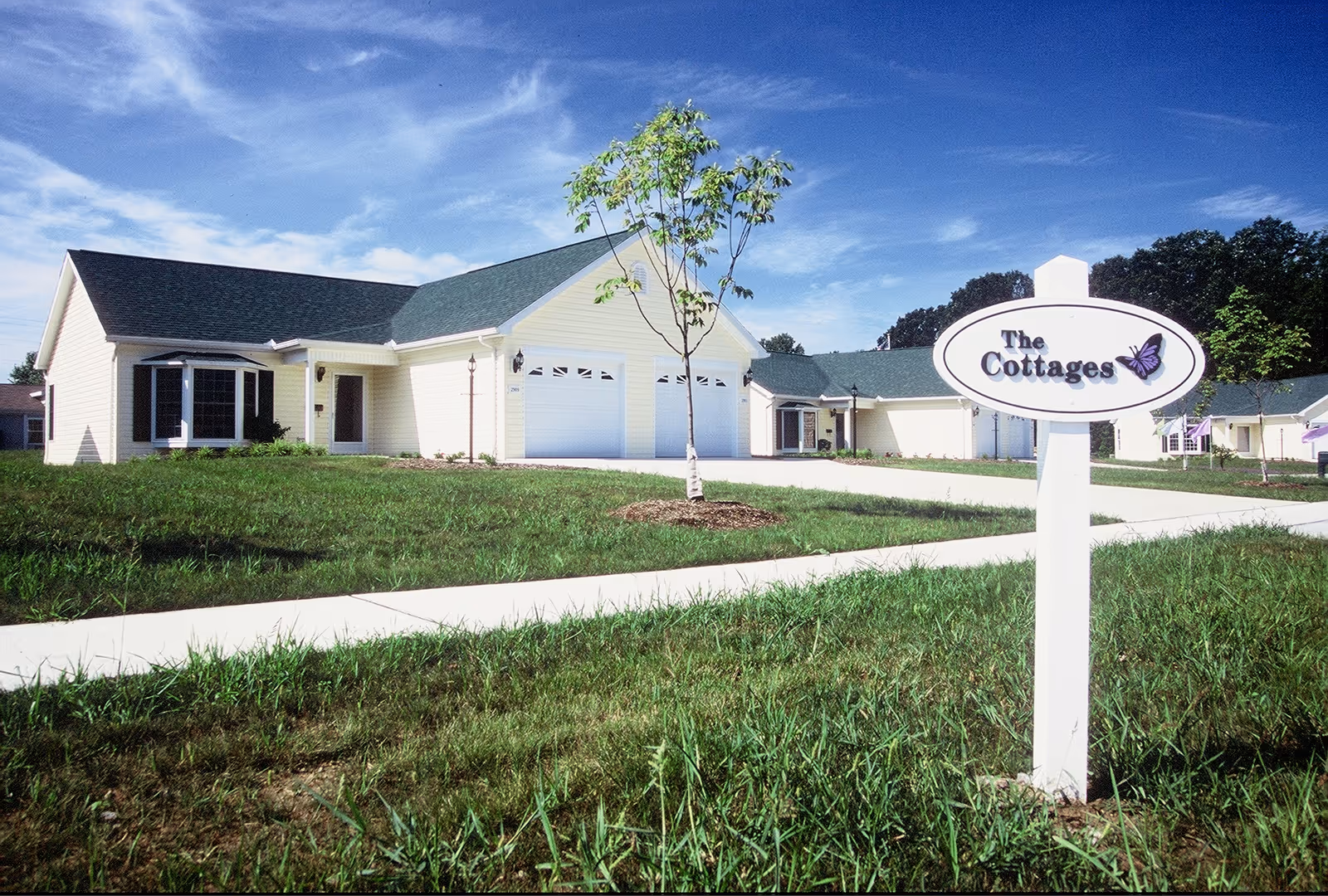 Single-story cottage-style senior living homes with a front lawn, sidewalk and a sign reading "The Cottages".