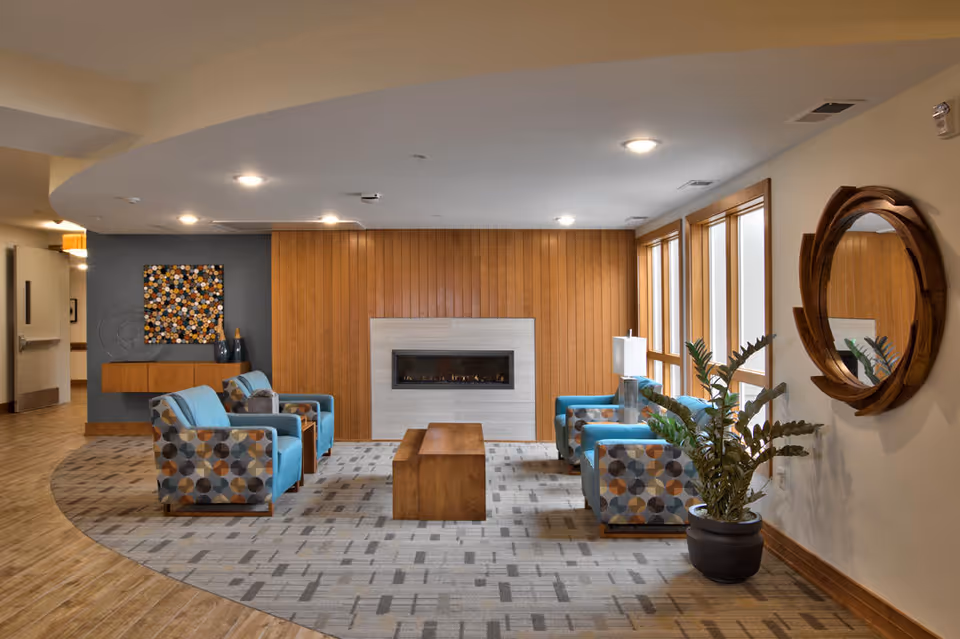 A cozy lounge area with four patterned armchairs arranged around a wooden coffee table in front of a modern fireplace set in a wooden paneled wall. Large windows on the right side let in natural light, and a round wooden-framed mirror hangs on the wall above a potted plant. The floor is a mix of wood and carpet with a geometric pattern, and there is a decorative wall art piece on the left wall.