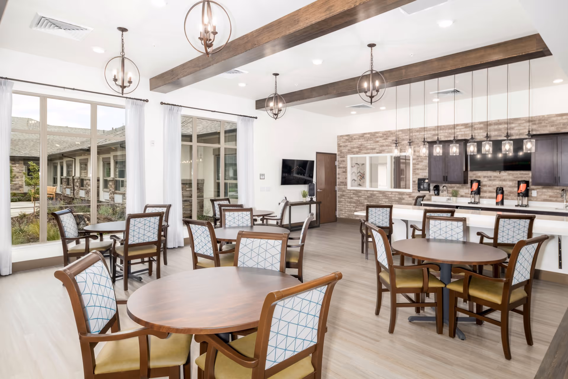 Bright and spacious dining room in a senior living facility with multiple round wooden tables and cushioned chairs featuring patterned backs. Large windows with white curtains allow natural light to fill the room. The ceiling has wooden beams and modern pendant lights. A counter area with beverage dispensers and a TV mounted on a brick accent wall are visible in the background.
