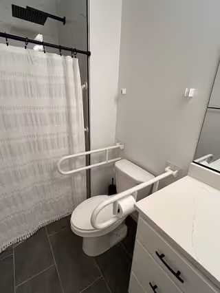 A modern bathroom featuring a white toilet with safety grab bars on both sides, a white shower curtain with a subtle pattern, a black showerhead, a white vanity with a marble countertop, and a large mirror above the sink. The floor is covered with dark gray tiles.