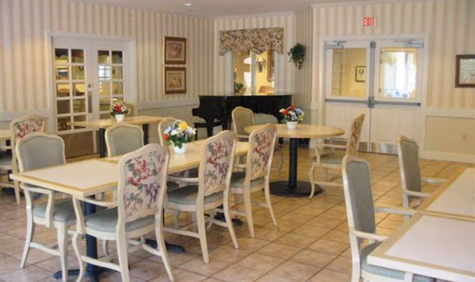 A dining room with several tables and chairs arranged neatly. The chairs have floral upholstery. There are small flower arrangements on the tables. The room has tiled flooring, striped wallpaper, and a black grand piano against the wall. Double doors with glass panels and an exit sign above are visible in the background.