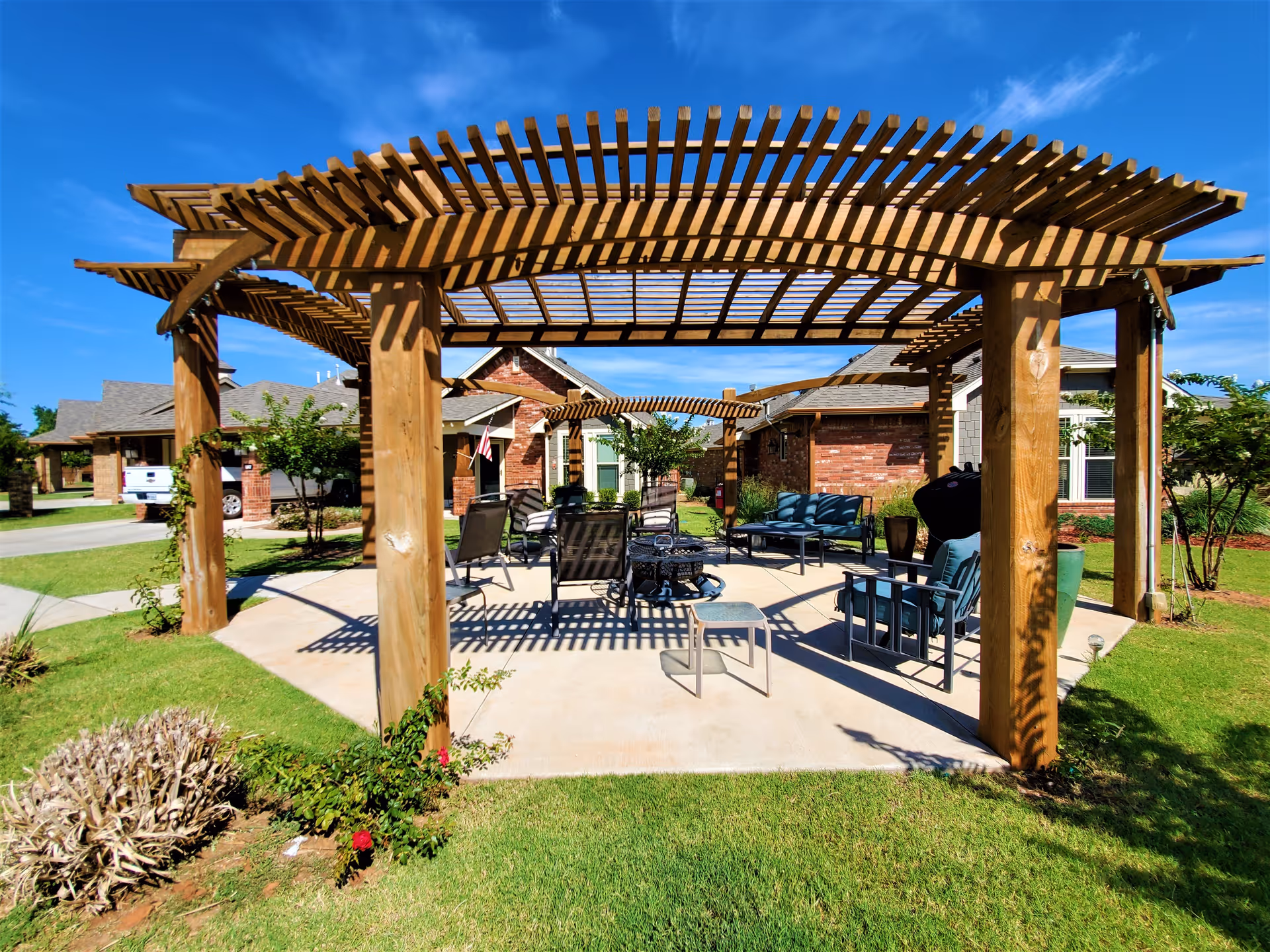 Outdoor seating area under wooden pergolas with chairs, a small table, and a fire pit on a concrete patio surrounded by grass and shrubs, with residential buildings in the background under a clear blue sky.