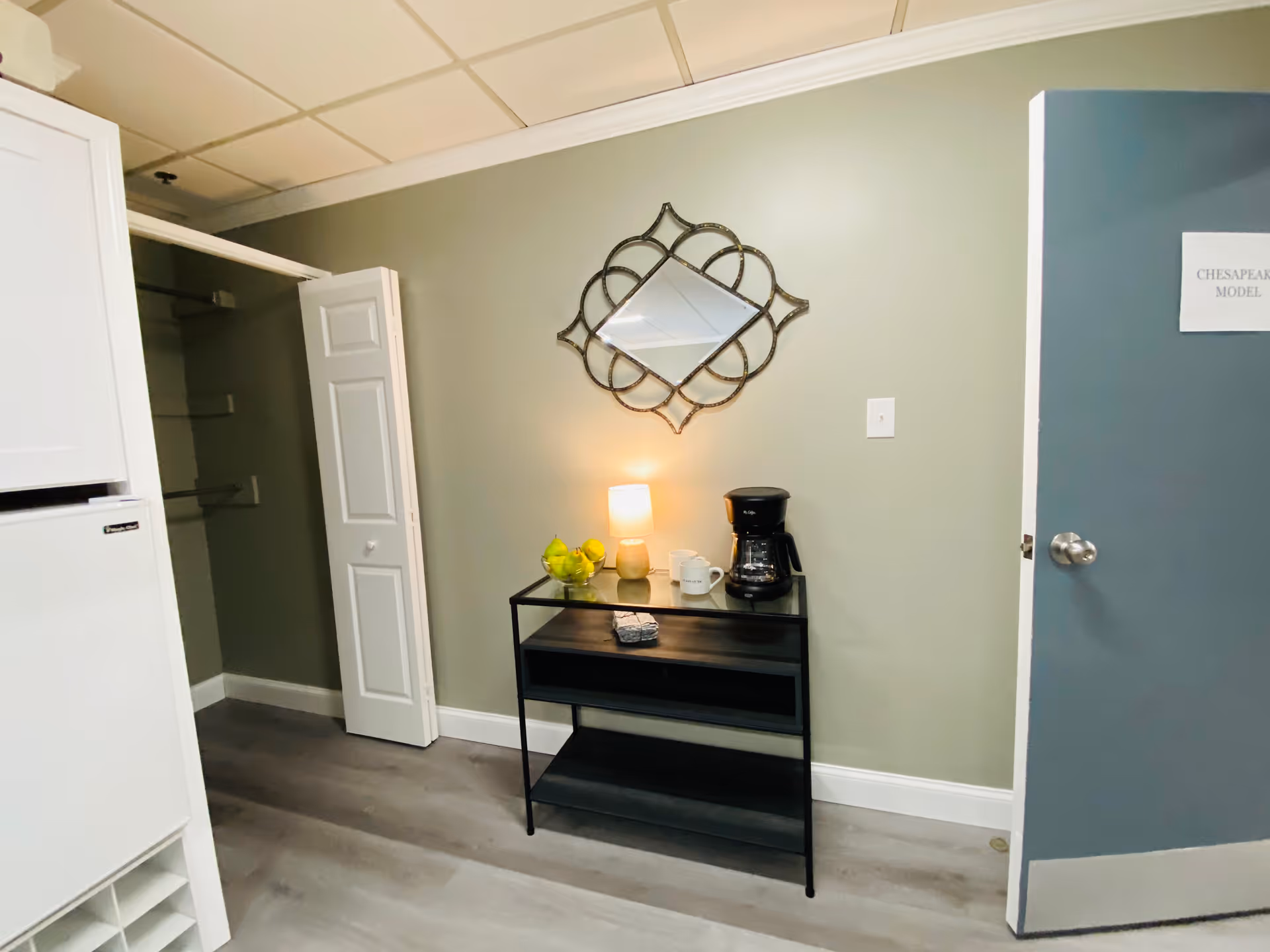 Interior room with light green walls and a gray door labeled 'Chesapeake Model'. A black metal table with a glass top holds a coffee maker, two mugs, a small lamp, and a bowl of green apples. Above the table is a decorative wall mirror with a geometric frame. To the left is a white closet with bi-fold doors and a white refrigerator partially visible.