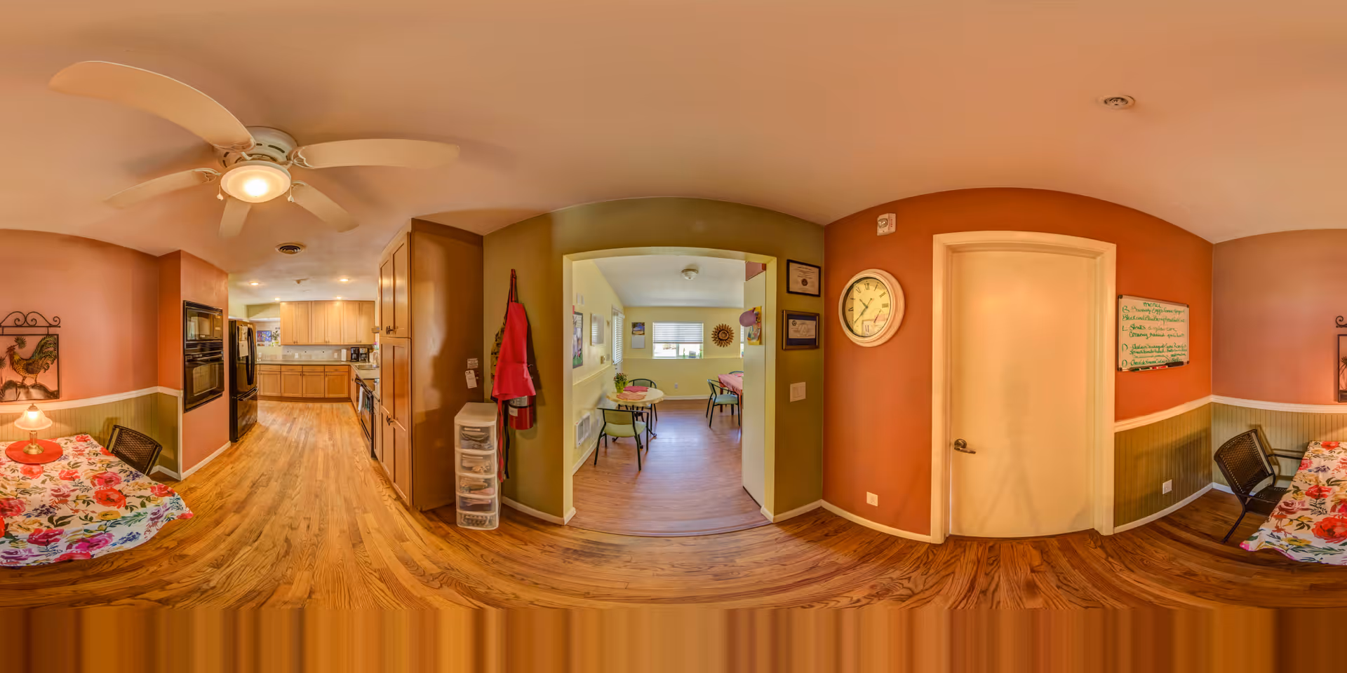 Panoramic view of an interior space in an assisted living facility showing a kitchen area with wooden cabinets and appliances, a dining area with tables covered in floral tablecloths, chairs, a ceiling fan, a clock on the wall, and a doorway leading to another room with more tables and chairs.