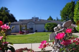 A single-story building with a gray roof and white walls surrounded by a well-maintained green lawn and colorful pink flowers in the foreground. There is a small statue of a fairy among the flowers, and trees are visible in the background under a clear blue sky.