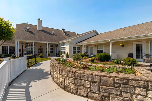 Outdoor courtyard area of a senior living facility with a stone planter filled with plants and flowers, surrounded by a concrete walkway and beige buildings with white trim and multiple doors and windows. There are chairs and hanging plants along the building walls.