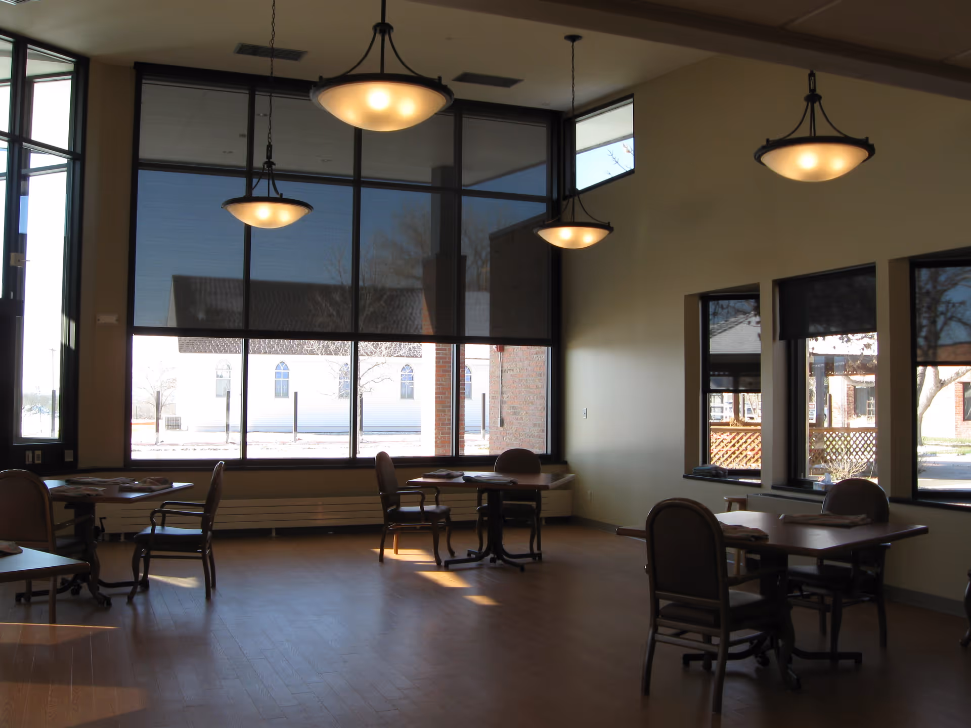 Sunlit common dining room with tables and chairs, hanging pendant lights, and large windows.