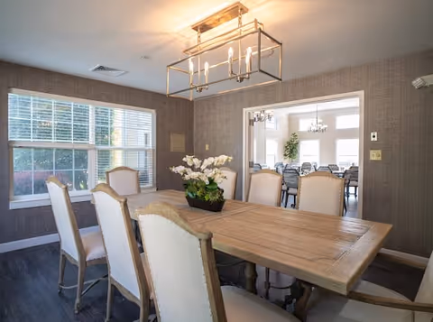 A dining room with a large wooden table surrounded by eight upholstered chairs. A rectangular chandelier with multiple lights hangs above the table. There is a window with blinds on the left wall, and an open doorway leads to another room with additional seating and large windows letting in natural light.
