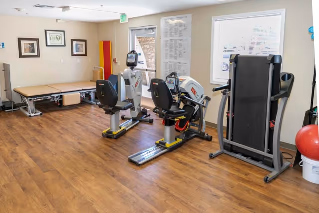A fitness room with exercise equipment including two recumbent stationary bikes, a treadmill, and a large exercise ball. There is a padded therapy table against the wall, framed pictures on the wall, and a window with blinds. The floor is wooden and the room is well-lit with natural light coming through the window and door.