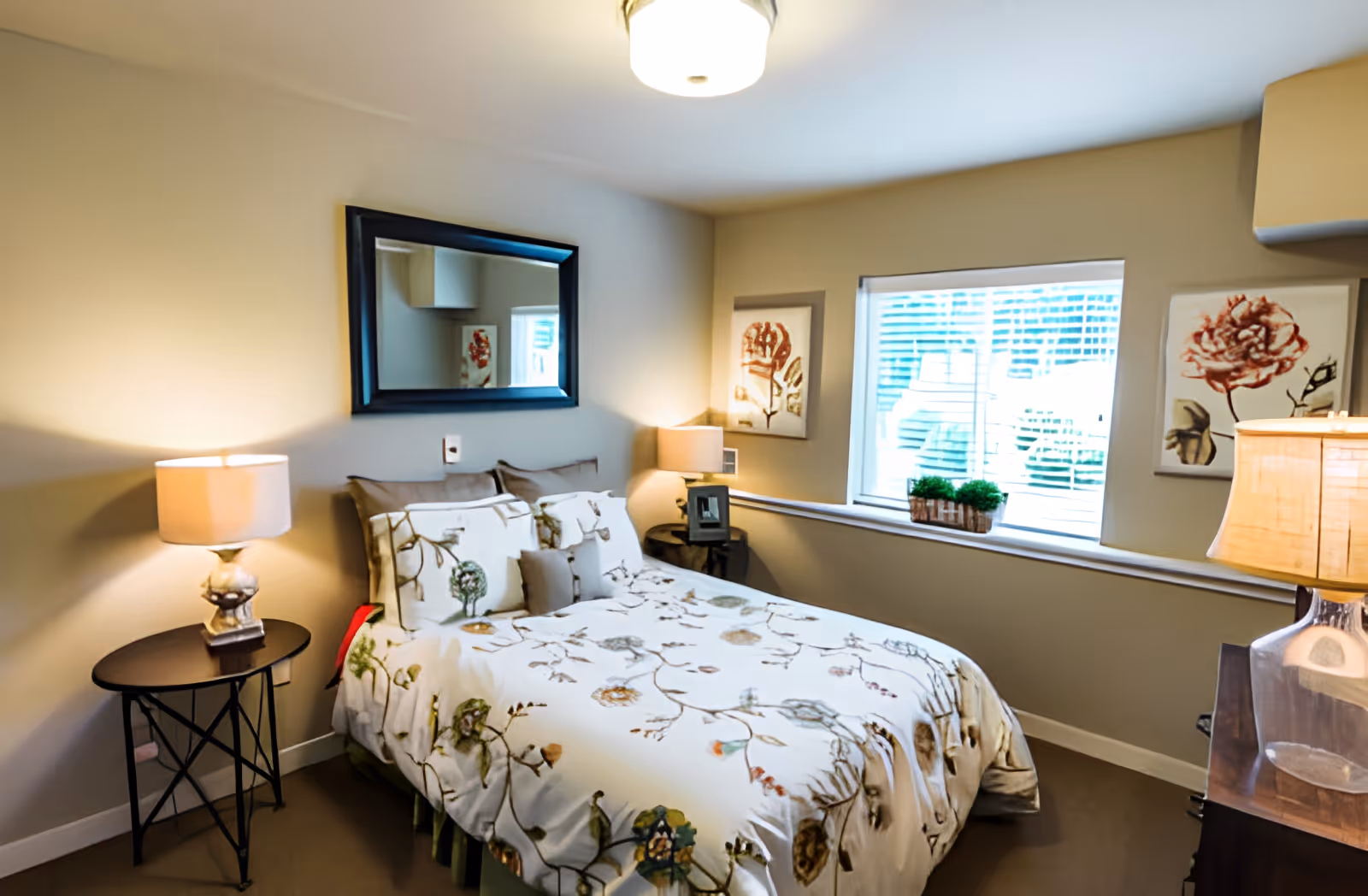 A cozy bedroom with a bed covered in a white floral-patterned comforter and multiple pillows. There are two bedside tables, each with a lamp, one on the left and one on the right side of the bed. Above the bed is a large rectangular mirror. The room has beige walls, a window with blinds, and two framed floral artworks on either side of the window. A small plant is placed on the window sill.