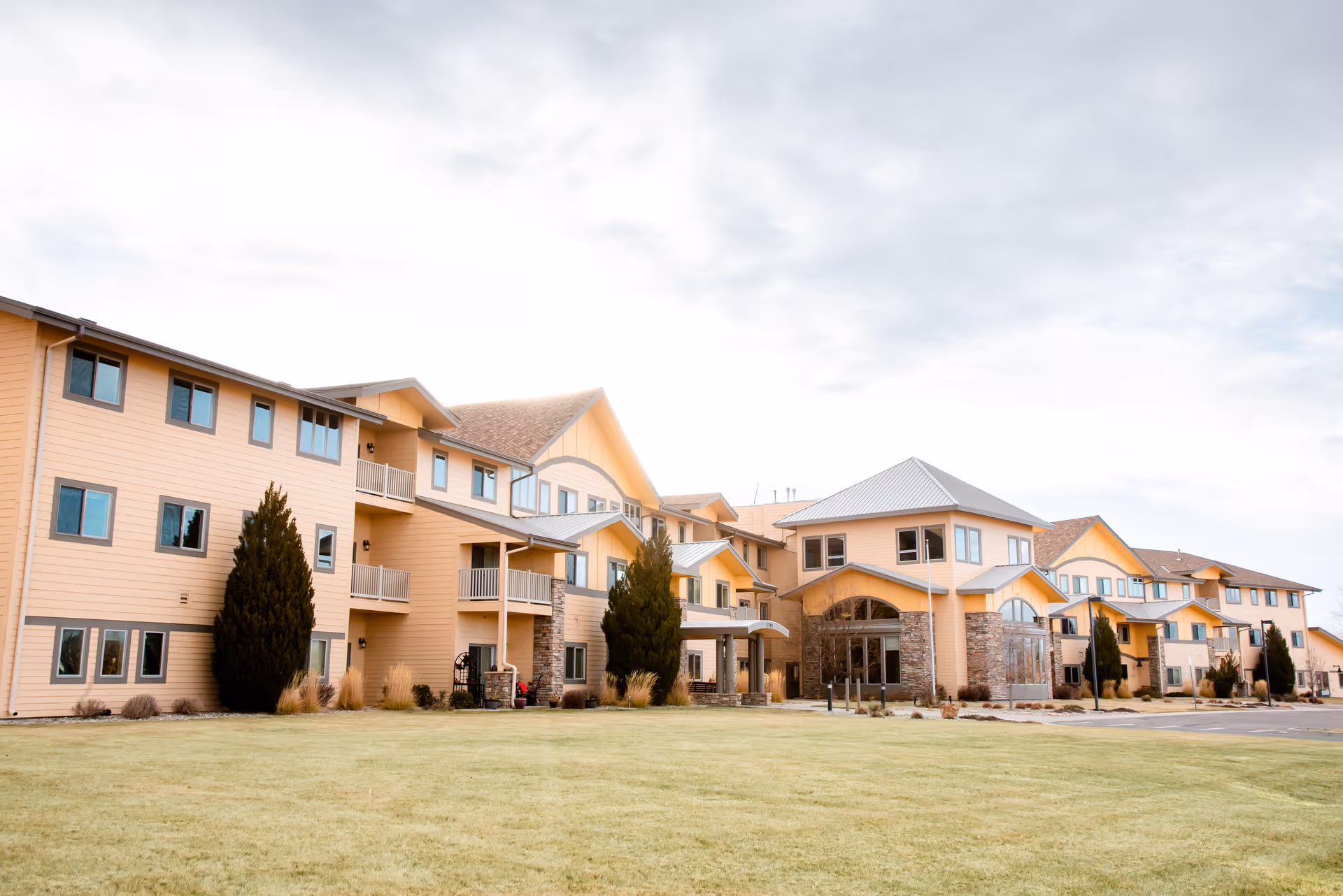 Front exterior of a multi-story beige senior living facility with balconies, entrance canopy, and a grassy lawn.