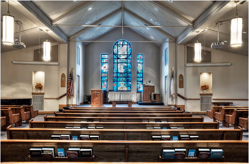 Interior view of a chapel with wooden pews facing an altar. The altar area features a large stained glass window with religious imagery, two wooden podiums, and flags on either side. The ceiling is vaulted with hanging pendant lights, and there are floral arrangements in wall niches on both sides.