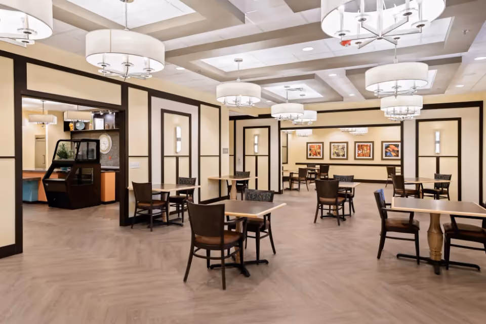 A spacious dining room with multiple square wooden tables and dark wooden chairs arranged neatly. The room features beige walls with dark trim, modern light fixtures hanging from the ceiling, and colorful framed artwork on the far wall. There is an open doorway leading to a kitchen or serving area with additional lighting and cabinetry.