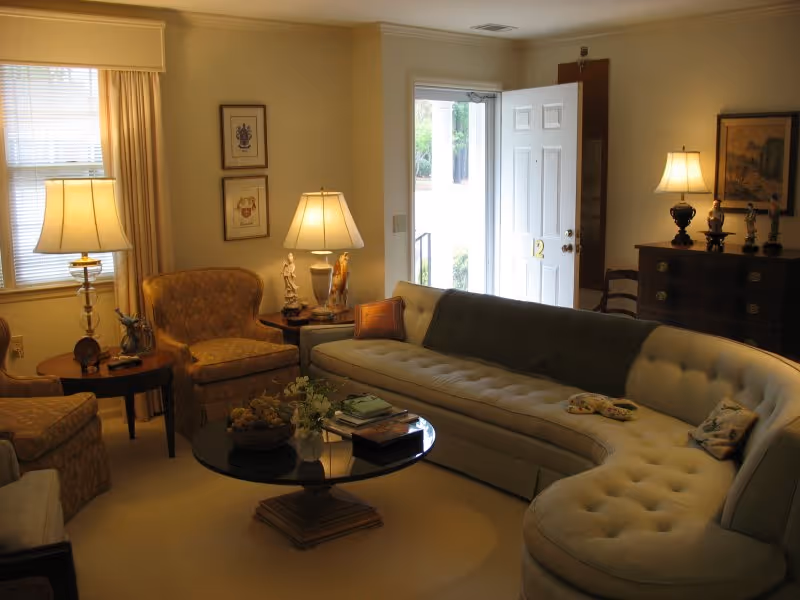A cozy living room with a large curved beige sofa, two upholstered armchairs, a round glass coffee table with books and decorative items, two table lamps on side tables, framed artwork on the walls, and an open door letting in natural light.