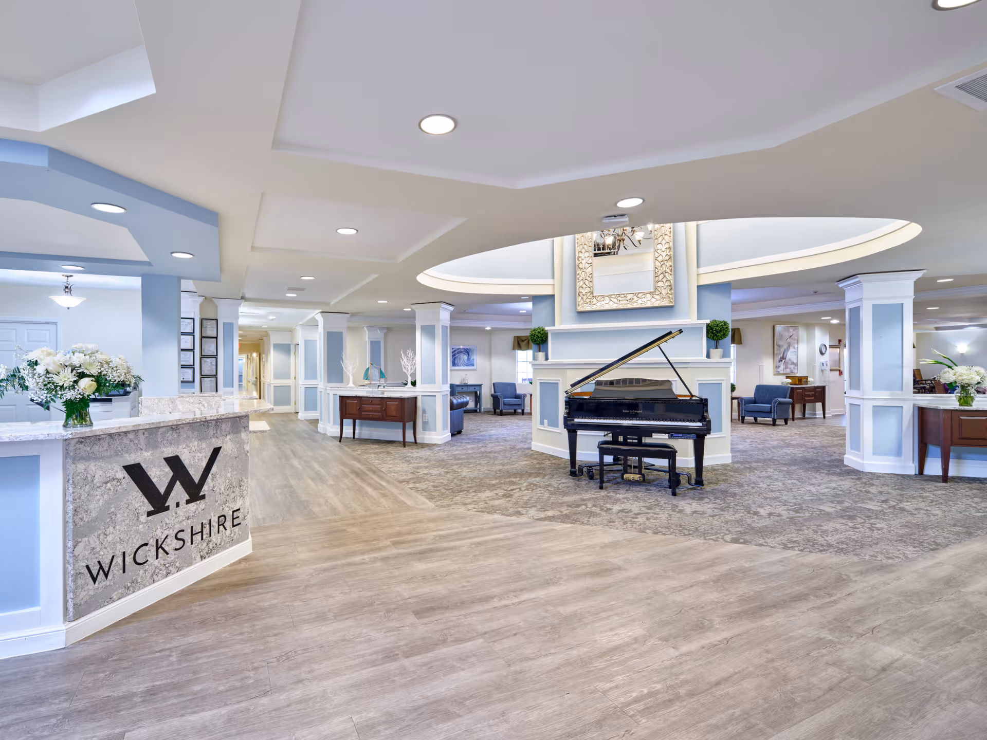 Spacious and well-lit senior living facility lobby with a grand piano in the center, a reception desk with the Wickshire logo, comfortable seating areas, decorative plants, and elegant lighting fixtures.