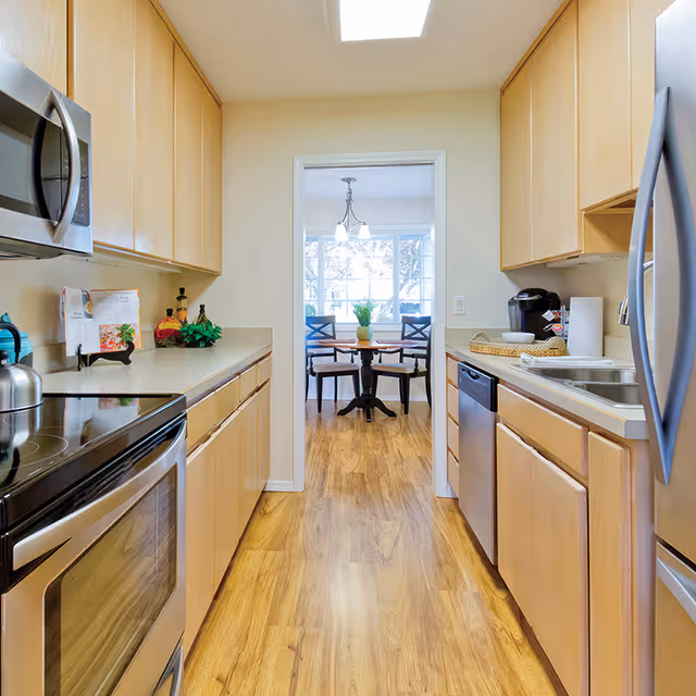 A bright kitchen with light wood cabinets on both sides, stainless steel appliances including a microwave, oven, dishwasher, and refrigerator. The kitchen has a light wood floor and a doorway leading to a dining area with a round table, four chairs, a potted plant centerpiece, and large windows letting in natural light.