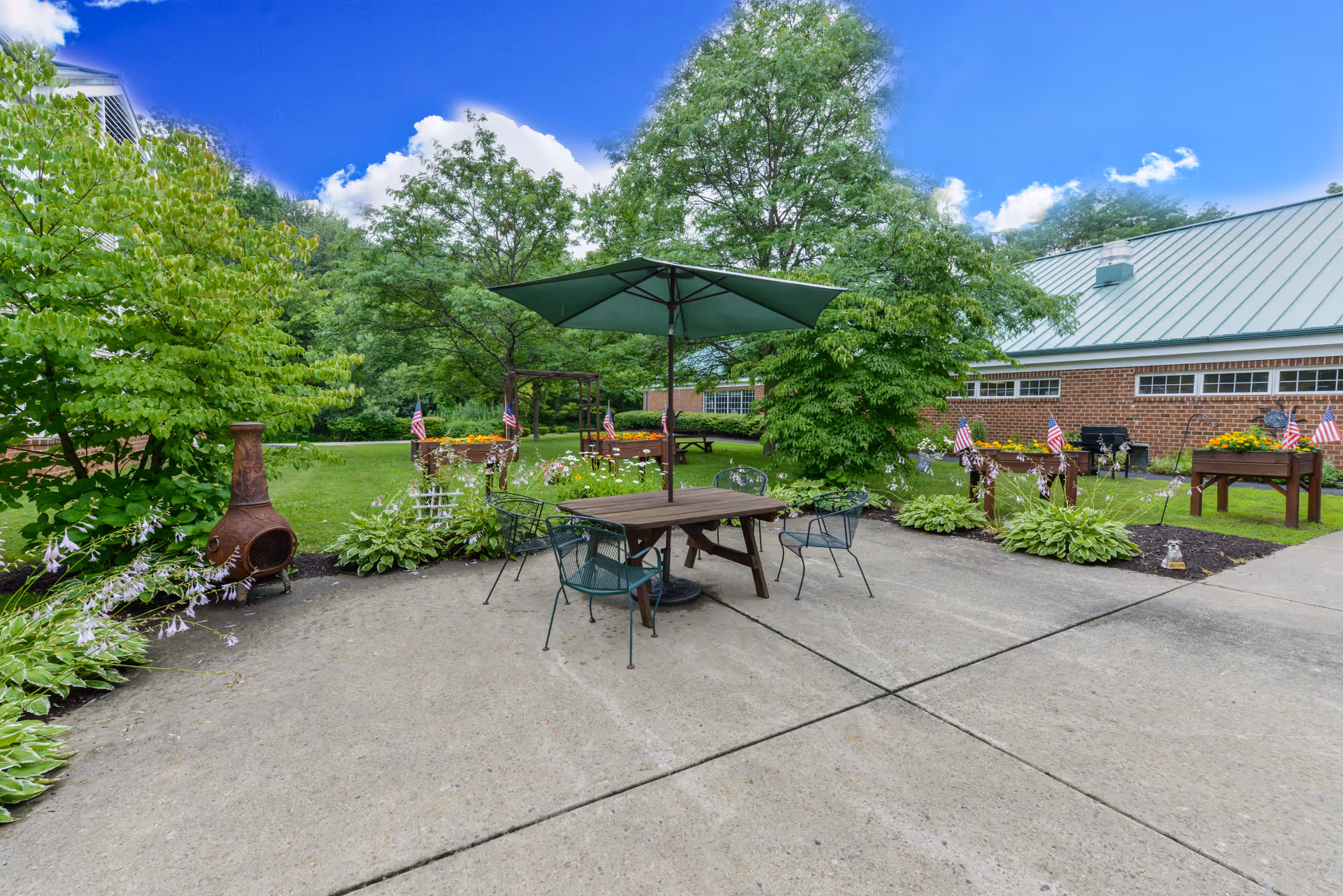 Outdoor patio area with a wooden table and four metal chairs under a large green umbrella. Surrounding the patio are lush green trees, plants, flower beds with American flags, and a brick building with a green metal roof in the background. The sky is clear with a few clouds.