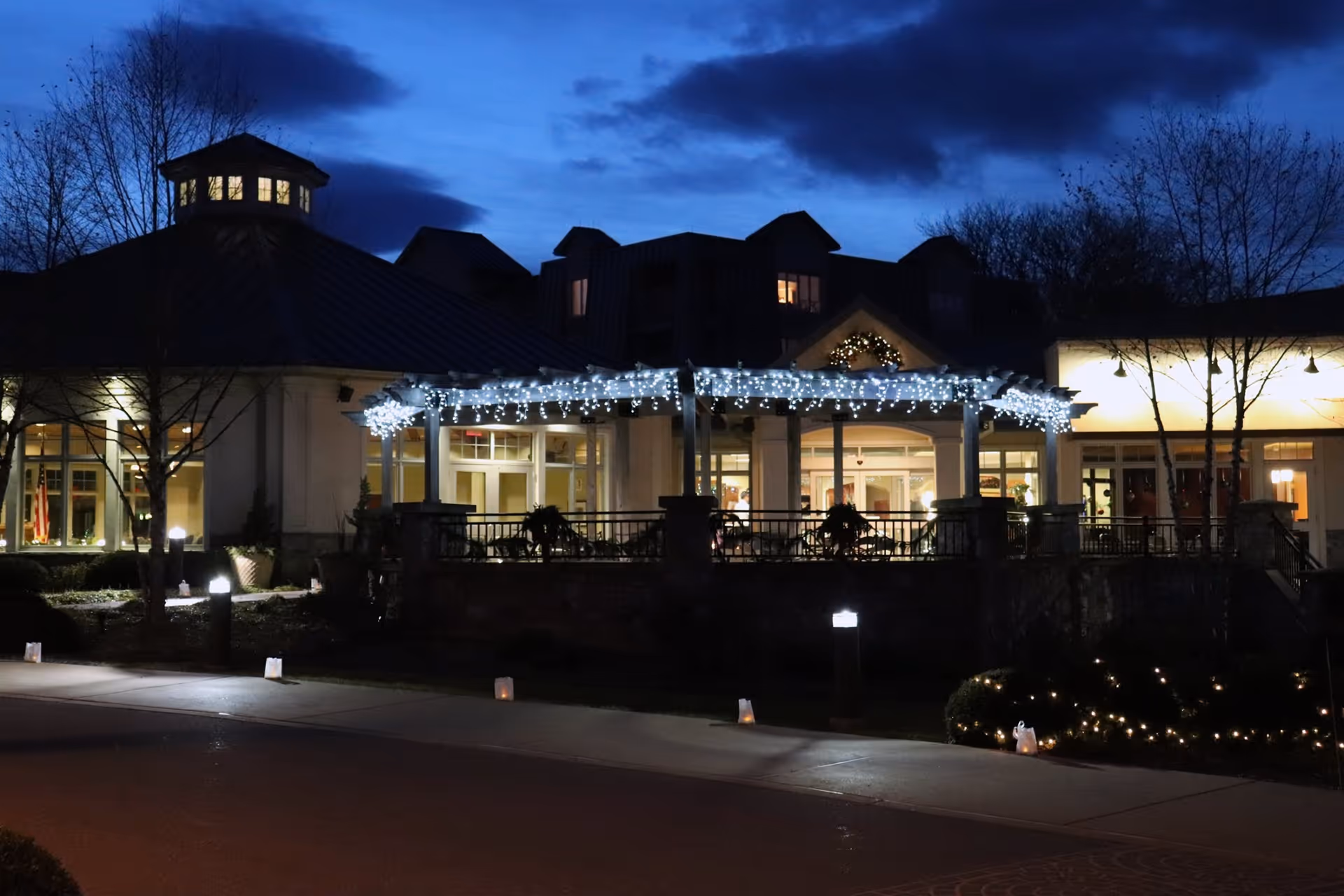 Exterior view of a senior living facility at dusk with decorative white string lights hanging from a pergola. The building has large windows with warm interior lighting and a dark blue sky with clouds in the background. There are leafless trees and small illuminated bags lining the sidewalk in front.